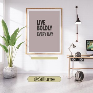 May include: A framed print with the text "LIVE BOLDLY EVERY DAY" in a modern interior setting. The artwork is in a wooden frame and hangs above a desk with a computer and lamp. A potted plant and fan are also visible.