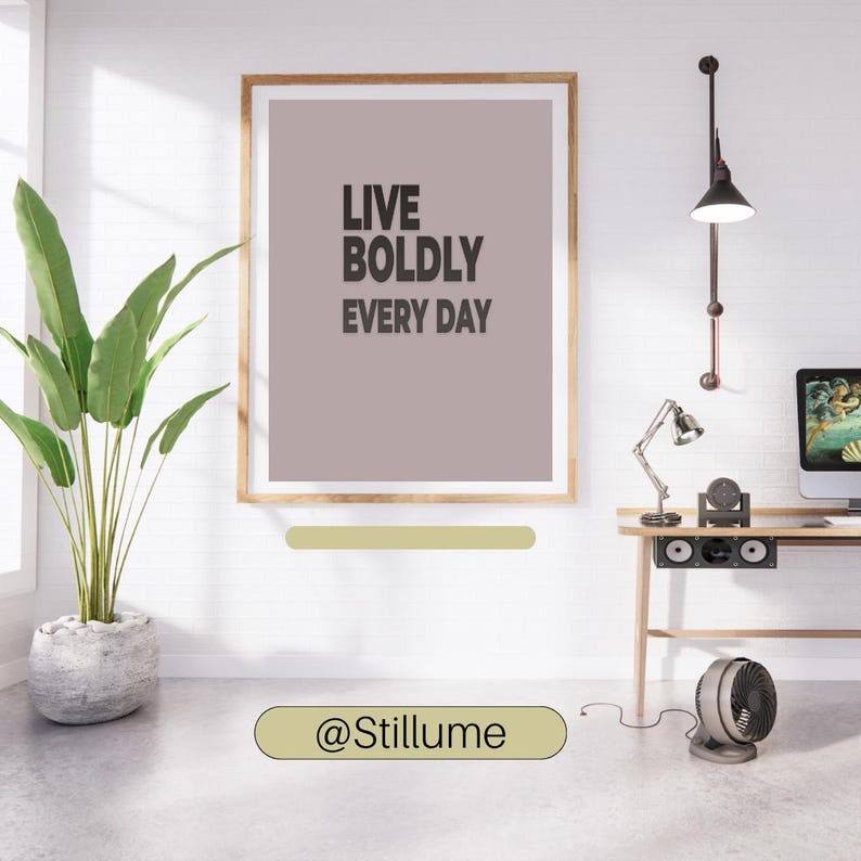 May include: A framed print with the text "LIVE BOLDLY EVERY DAY" in a modern interior setting. The artwork is in a wooden frame and hangs above a desk with a computer and lamp. A potted plant and fan are also visible.