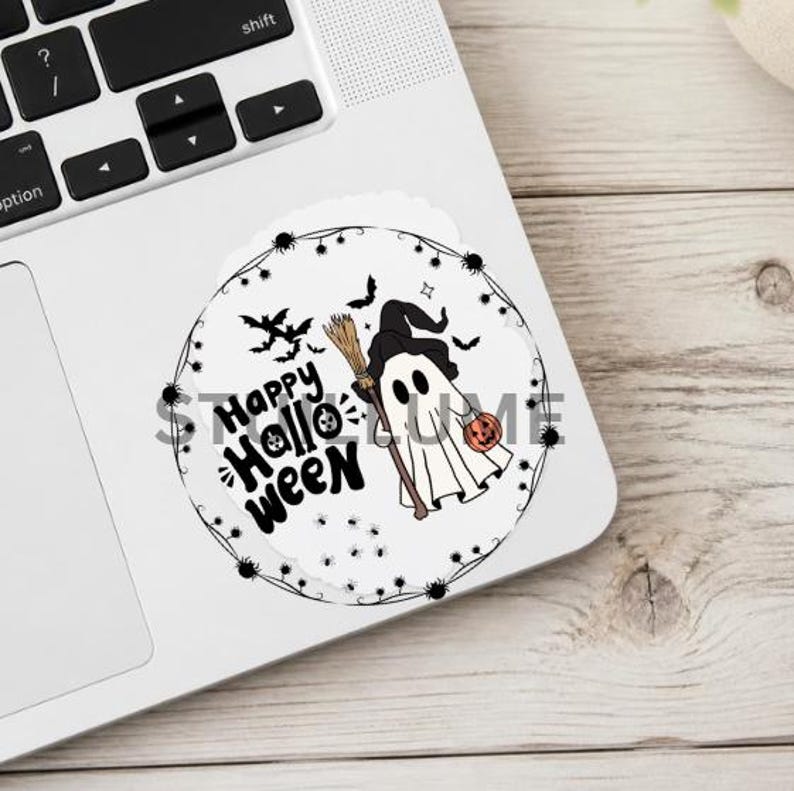 May include: A circular sticker featuring a white ghost wearing a black witch hat, holding a brown broom and an orange pumpkin bucket. The text reads Happy Halloween in black script, surrounded by a thin border of small black spiders and webs.
