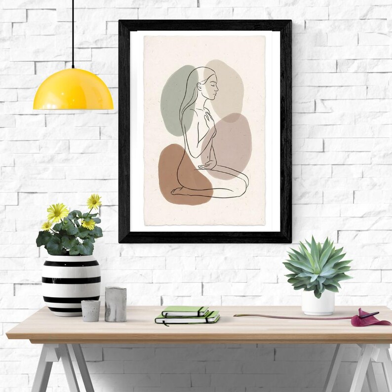 Pu&ograve; includere: Una stampa artistica da parete incorniciata che presenta un disegno a linee nere di una persona inginocchiata di profilo. La figura &egrave; sovrapposta a forme astratte dai toni tenui verde salvia, beige e terracotta su uno sfondo crema chiaro.