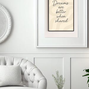 May include: A framed print with the text "Dreams are better when shared" in a black frame. The background is a light beige color with wavy lines. The print is displayed on a white wall above a white chair.