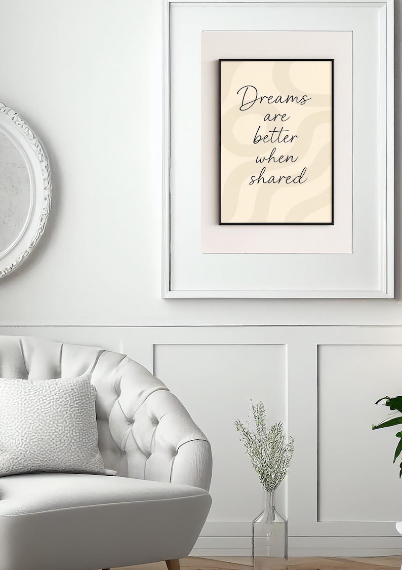 May include: A framed print with the text "Dreams are better when shared" in a black frame. The background is a light beige color with wavy lines. The print is displayed on a white wall above a white chair.