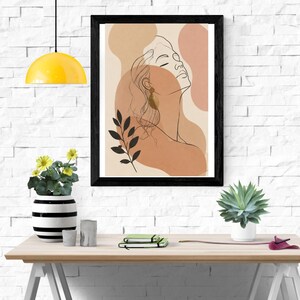 May include: A framed wall art print featuring a black line drawing of a person's profile against abstract shapes in shades of beige, tan, and terracotta. The artwork includes a stylized black leaf branch and is displayed in a black frame.