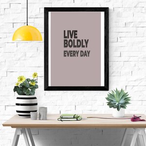 May include: A framed print with the words "LIVE BOLDLY EVERY DAY" in black text. The print is displayed on a wooden desk with a yellow lamp, a black and white striped vase with yellow flowers, and a small succulent in a white pot.