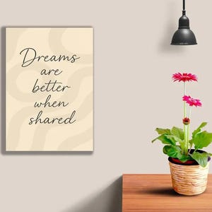 May include: A framed print with the text "Dreams are better when shared" in a cursive font. A potted gerbera daisy with bright pink flowers sits on a wooden table. A black pendant light hangs above.