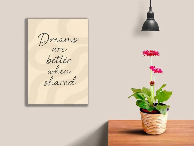 May include: A framed print with the text "Dreams are better when shared" in a cursive font. A potted gerbera daisy with bright pink flowers sits on a wooden table. A black pendant light hangs above.