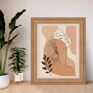 May include: A framed wall art print featuring a black line drawing of a person in profile with closed eyes, set against abstract shapes in shades of beige, tan, and terracotta, accented by a black leaf illustration and a gold-toned earring.