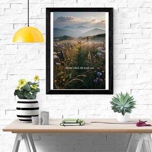 May include: A framed wall art print featuring a landscape of a wildflower meadow at sunset with a butterfly in the center and the text Bloom where life leads you. The print is displayed in a black frame on a white brick wall above a wooden desk.