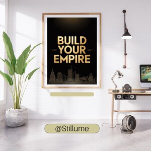 May include: A framed wall art print featuring a black background with the gold text BUILD YOUR EMPIRE centered above a gold outline of a city skyline. The print is displayed in a light wood frame on a white wall, positioned above a desk area.