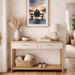 May include: A watercolour painting in a wooden frame shows a couple on a bench at sunset. The artwork hangs above a light wood console table with two drawers, a woven basket, and a lamp. A beige rug and vase with dried plants complete the decor.