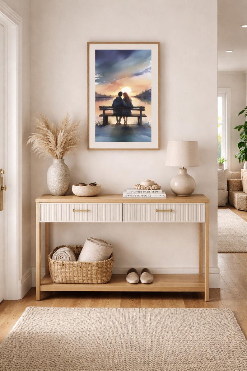 May include: A watercolour painting in a wooden frame shows a couple on a bench at sunset. The artwork hangs above a light wood console table with two drawers, a woven basket, and a lamp. A beige rug and vase with dried plants complete the decor.