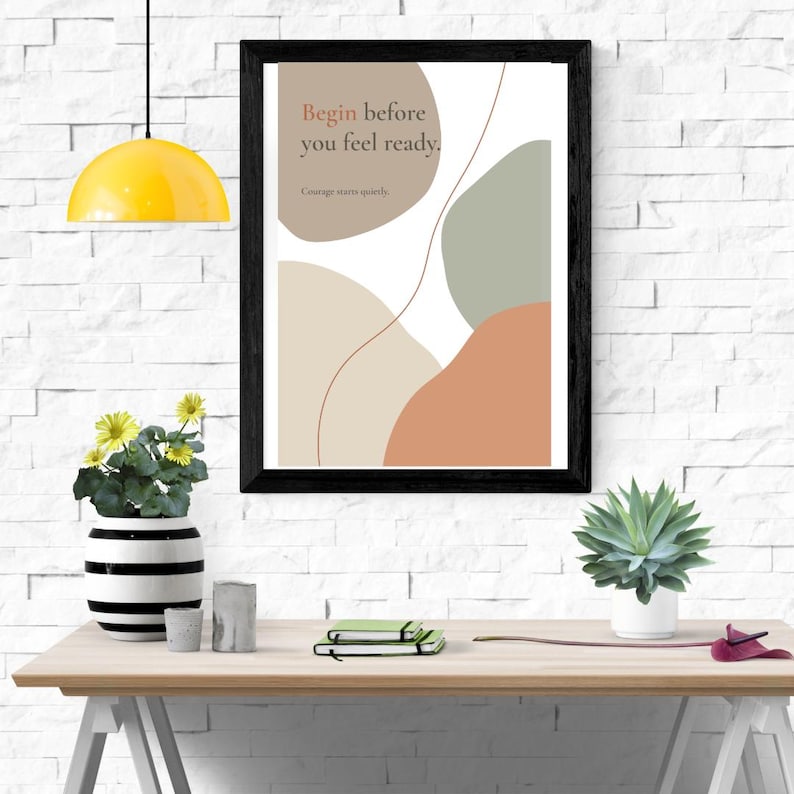 K&ouml;nnte beinhalten: Ein gerahmter Wandkunstdruck mit abstrakten organischen Formen in gedeckten T&ouml;nen von Beige, Salbeigr&uuml;n und Terrakotta. Der Text lautet: "Begin before you feel ready. Courage starts quietly." Der Druck befindet sich in einem schwarzen Rahmen.