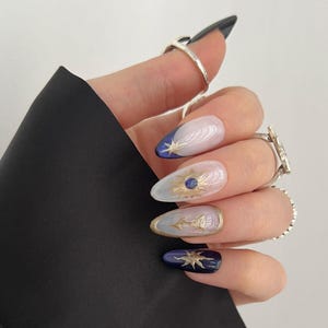 May include: A hand with long, almond-shaped nails. The nails are painted with a celestial design in gold, blue, and white. The thumb nail is black. The hand is wearing several silver rings and is emerging from a black sleeve.