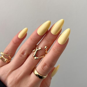 Könnte beinhalten: Eine Hand mit langen, mandelförmigen Nägeln, die in einem blassen Gelb lackiert sind. Die Hand ist mit drei goldenen Ringen unterschiedlichen Designs geschmückt: ein breites Band, ein skulpturales Stück und ein Doppelring. Nägel und Ringe erzeugen eine stilvolle, moderne Ästhetik.