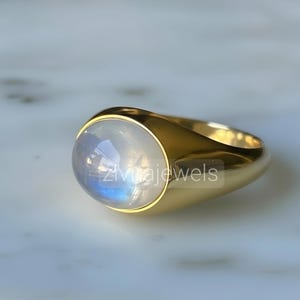Puede incluir: Un anillo de sello de oro con una piedra lunar cabujón ovalada y lisa, de aspecto translúcido y lechoso con un sutil brillo azul. La banda es de oro pulido con un diseño sólido y cónico que sujeta la piedra preciosa de forma segura.