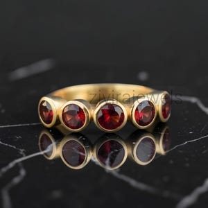 Peut inclure: Une bague en or ornée de cinq pierres précieuses grenat rondes et rouge foncé, serties dans un style clos. L'anneau présente une finition lisse et polie, et le texte zivirajewels est visible au centre. Le design affiche une disposition symétrique.