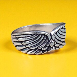 Op de afbeelding: Een zilverkleurige ring met een vleugelontwerp. De ring heeft gedetailleerde veertjes en staat tegen een heldergele achtergrond. Het vleugelontwerp bevindt zich aan de voorkant van de ring.