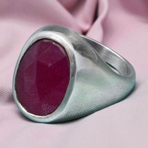 May include: A silver ring featuring a large, faceted, oval-shaped, deep red gemstone. The ring is set against a light pink fabric background, contrasting the cool metal with the warm gemstone.
