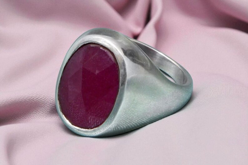 May include: A silver ring featuring a large, faceted, oval-shaped, deep red gemstone. The ring is set against a light pink fabric background, contrasting the cool metal with the warm gemstone.