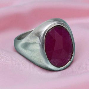 May include: A silver ring featuring a large, oval, faceted ruby gemstone. The ring has a wide band and a brushed metal finish. The ruby is a deep, rich red colour. The ring is set against a pink fabric background.