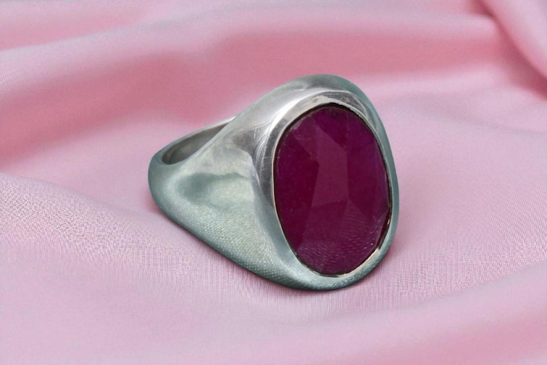 May include: A silver ring featuring a large, oval, faceted ruby gemstone. The ring has a wide band and a brushed metal finish. The ruby is a deep, rich red colour. The ring is set against a pink fabric background.