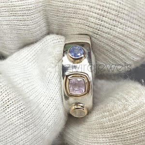 Puede incluir: Un anillo de banda de plata con tres piedras preciosas alineadas verticalmente. La piedra superior es una gema redonda azul claro, el centro es una gema cuadrada rosa claro en un engaste de oro y la inferior es una gema redonda transparente.