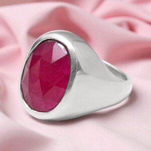 May include: A silver ring featuring a large, oval-cut ruby gemstone. The ring is set against a pink fabric, accentuating the deep red hue of the stone. The ring's band has a smooth, polished finish.