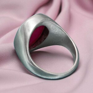 May include: A silver ring featuring a large, oval, magenta gemstone. The ring is set against a soft, pink fabric, accentuating the contrast between the metal and the stone. The ring's band is thick and smooth.