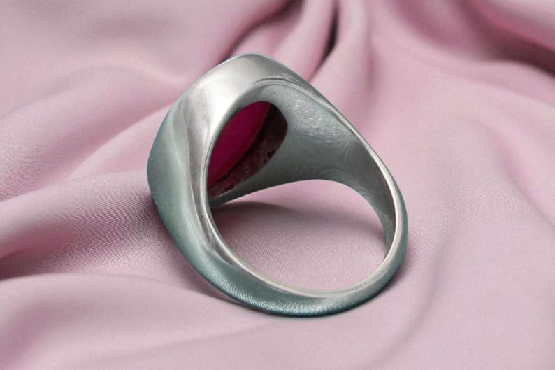 May include: A silver ring featuring a large, oval, magenta gemstone. The ring is set against a soft, pink fabric, accentuating the contrast between the metal and the stone. The ring's band is thick and smooth.