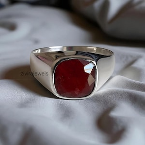 Pode incluir: Um anel de prata com uma grande pedra preciosa vermelha escura de corte quadrado. O anel tem uma banda larga e um acabamento polido. A pedra preciosa é facetada, captando a luz e exibindo uma cor rica e vibrante. O anel é exibido em uma superfície branca texturizada.
