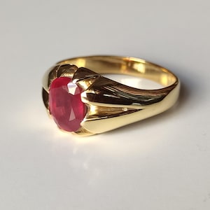 Può includere: Un anello d'oro con una grande gemma di rubino taglio ovale. L'anello ha un design classico con una fascia in oro lucido e un'incastonatura che tiene saldamente la vibrante pietra rossa. L'anello è esposto su una superficie bianca.