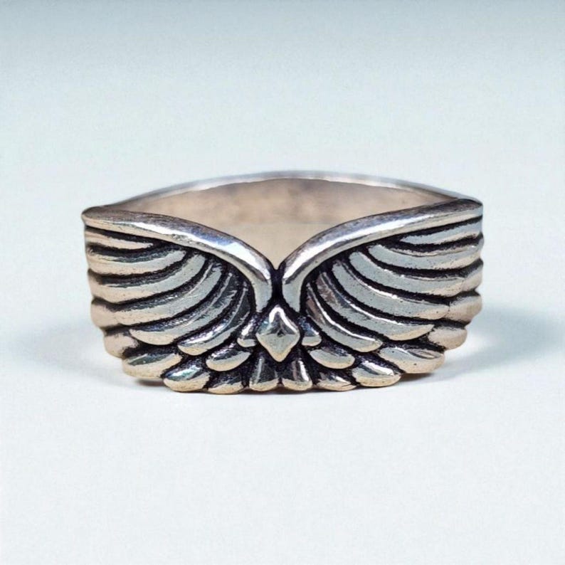 Op de afbeelding: Een zilveren ring met een gedetailleerd vleugelontwerp. De ring heeft een centraal punt en gelaagde veerd details, waardoor een textuur en dimensionale uitstraling ontstaat. De ring staat tegen een effen witte achtergrond.