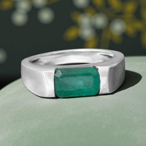 Pode incluir: Um anel de prata com uma pedra preciosa retangular verde esmeralda. O anel tem uma banda plana de metal escovado e uma moldura de bisel. O anel é exibido em uma superfície verde clara.