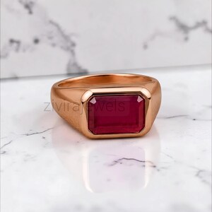 May include: A rose gold signet ring featuring a rectangular, emerald-cut deep red gemstone set in the centre. The band has a smooth, polished finish with a textured side detail. The text zivirajewels is visible across the centre of the ring.