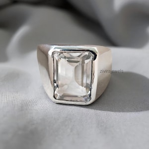 Puede incluir: Un anillo de plata con una piedra preciosa rectangular y transparente. El anillo tiene una banda ancha y un acabado pulido. La piedra preciosa está engastada en un bisel. El texto "zivirajewels" es visible en el lateral.
