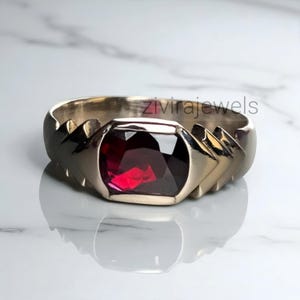 Pode incluir: Um anel de prata com uma grande pedra retangular vermelha escura. O anel tem um design único com detalhes angulares nas laterais. O nome da marca "zivirajewels" é visível.