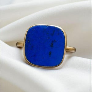 Pode incluir: Um anel dourado com uma grande pedra preciosa quadrada azul escura. A pedra preciosa tem uma forma ligeiramente arredondada e está engastada numa banda simples. O anel é exibido numa superfície branca texturizada.