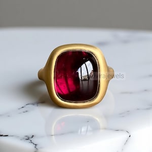 May include: A gold-toned ring featuring a large, square-cut, deep red gemstone. The ring has a smooth, matte finish and a substantial band. The gemstone is set in a bezel setting, enhancing its rich color. The ring is displayed on a white marble surface.