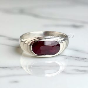May include: A silver ring featuring a large, rectangular, deep red gemstone. The ring has a smooth, polished band and a bezel setting that securely holds the stone. The gemstone reflects light, creating a vibrant appearance. The ring is displayed on a marble surface.
