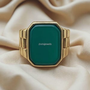 Può includere: Un anello dorato con una grande pietra rettangolare verde smeraldo. La fascia dell'anello ha un design segmentato. Il testo "zivirajewels" è stampato sulla pietra. L'anello è esposto su un tessuto beige.