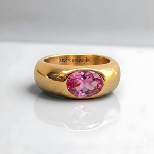 May include: A polished gold band ring featuring a central oval-cut pink gemstone. The interior of the band is engraved with the text zivirajewels. The ring has a smooth, rounded profile and a reflective metallic finish with a vibrant pink stone.