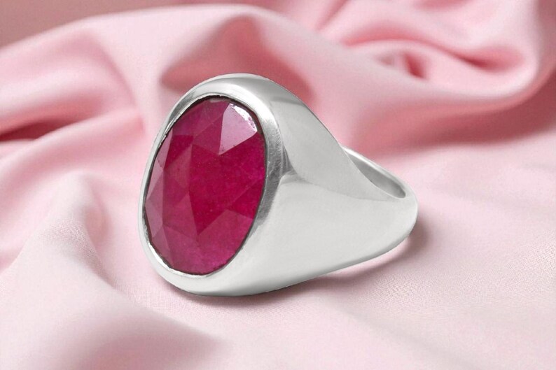May include: A silver ring featuring a large, oval-cut ruby gemstone. The ring is set against a pink fabric, accentuating the deep red hue of the stone. The ring's band has a smooth, polished finish.