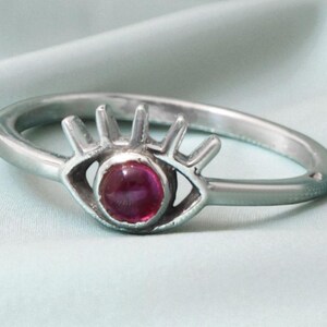May include: A silver-colored ring featuring an eye design with a central, round, deep red stone. The eye has a stylized, open shape with radiating lines above, set against a soft, light blue background.