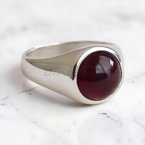 May include: A polished silver signet ring featuring a smooth, round, deep burgundy cabochon gemstone set in the centre. The metal band has a high-shine finish and a solid, tapered design. The text zivirajewels is visible on the ring surface.