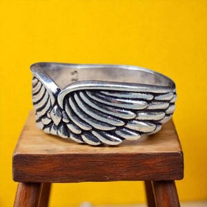 Op de afbeelding: Een zilverkleurige ring met een gedetailleerd vleugelontwerp. De ring, tegen een gele achtergrond, staat op een kleine houten kruk. Het vleugelontwerp heeft ingewikkelde veerdetaillering, wat een textuur cre&euml;ert. De band van de ring is zichtbaar, met een glad, gepolijst oppervlak.