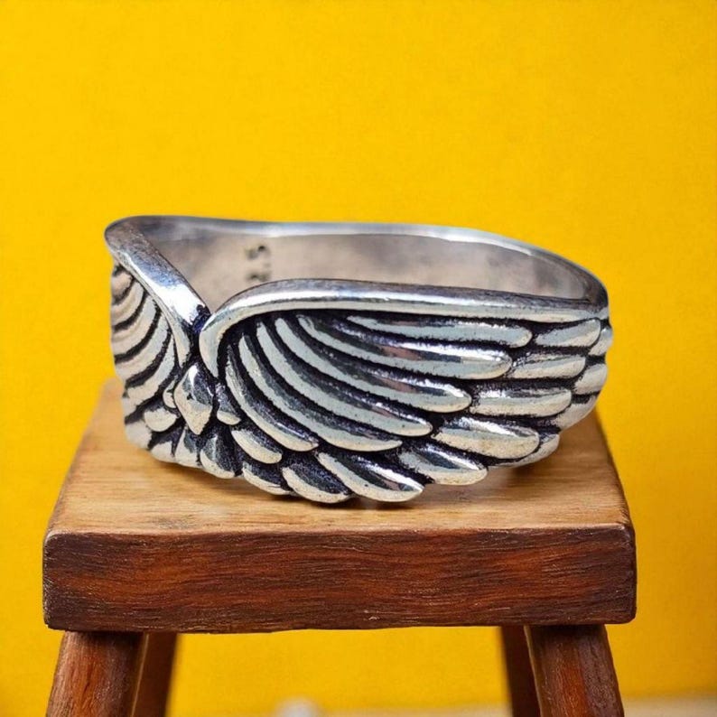 Op de afbeelding: Een zilverkleurige ring met een gedetailleerd vleugelontwerp. De ring, tegen een gele achtergrond, staat op een kleine houten kruk. Het vleugelontwerp heeft ingewikkelde veerdetaillering, wat een textuur cre&euml;ert. De band van de ring is zichtbaar, met een glad, gepolijst oppervlak.