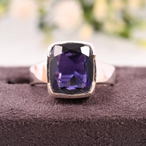 May include: A silver ring featuring a large, square-cut, deep purple gemstone. The gemstone is set in a silver bezel, and the band is a simple, polished silver. The ring is displayed on a dark purple, textured surface.