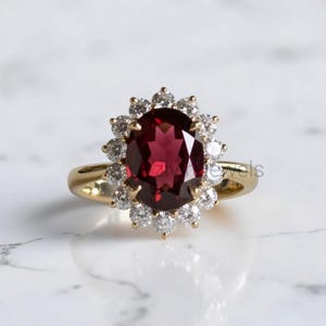 Peut inclure: Une bague en or avec une grande pierre grenat rouge foncé de taille ovale au centre, entourée d'un halo de petits diamants blancs ronds et clairs. L'anneau est en or jaune poli, formant un design en grappe classique et contrasté.