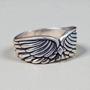 Op de afbeelding: Een zilverkleurige ring met een gedetailleerd vleugelontwerp. De ring heeft twee vleugels die zich om de band wikkelen, waarbij elke veer zorgvuldig is gesneden. De ring is waarschijnlijk gemaakt van sterling zilver of een vergelijkbaar metaal.