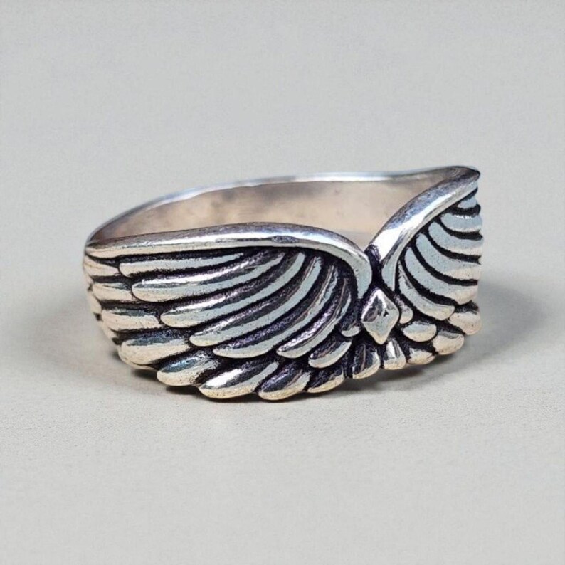 Op de afbeelding: Een zilverkleurige ring met een gedetailleerd vleugelontwerp. De ring heeft twee vleugels die zich om de band wikkelen, waarbij elke veer zorgvuldig is gesneden. De ring is waarschijnlijk gemaakt van sterling zilver of een vergelijkbaar metaal.